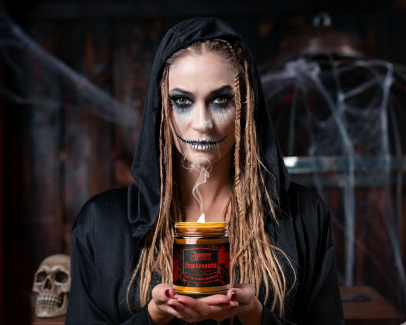 Person in black hood holding a candle with a Halloween-themed label, surrounded by spider webs and a skull.